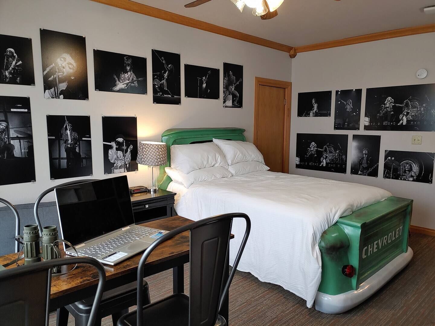 A hotel room with a bed, a desk with a laptop, a green Chevrolet toy car, and multiple black-and-white band posters on the walls.