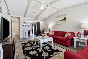 A living room with a red sofa, a coffee table, and a TV. There is a kitchen in the background with a dining area.