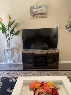 Ein Wohnzimmer mit Flachbildfernseher auf einem Holzboden, einer Topfpflanze und einer gerahmten Blumenkomposition auf einem Couchtisch.