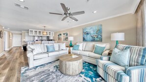 A spacious living room with a large sofa, a striped armchair, and a coffee table. There is a ceiling fan, a kitchen in the background, and artwork on the walls. - Stunning Beachfront Condo w/ Free Beach Service~ Crystal Dunes 405 (Destin)