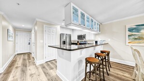 A modern kitchen with white cabinetry, a stainless steel refrigerator, a microwave, and a granite countertop. There are three wicker bar stools and a painting of a seascape on the wall. - Stunning Beachfront Condo w/ Free Beach Service~ Crystal Dunes 405 (Destin)