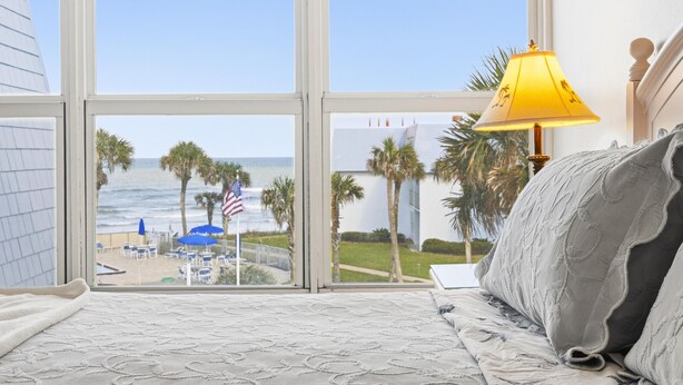 Ein Schlafzimmer mit großem Fenster mit Blick auf einen Strand, einem Bett mit gemusterten Bettwäsche und einer gelben Lampe.