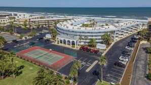 Luftaufnahme eines Küstenhotels mit Tennisplatz, Parkplatz und Meerblick.