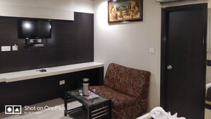 Een moderne woonkamer met een flatscreen televisie aan een donkere muur, een bank met een bloemenprint, een glazen salontafel met een roestvrijstalen waterfles, en een airconditioning.