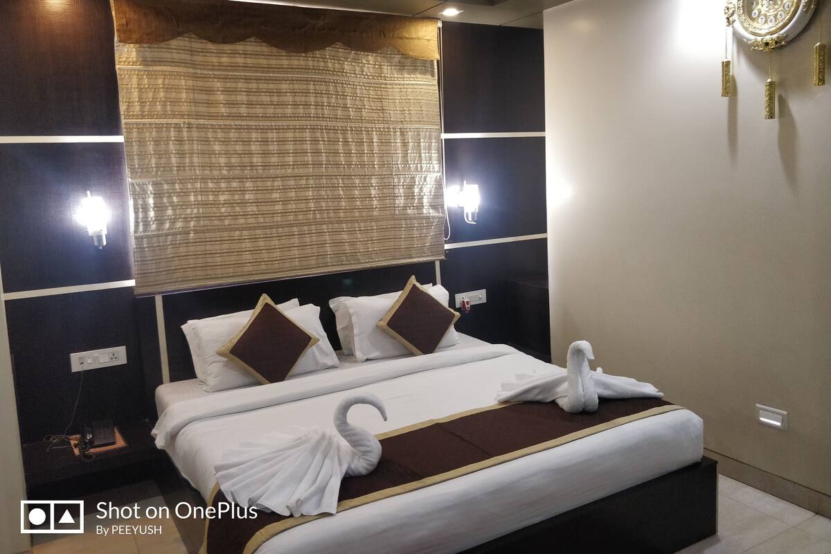 Quarto de hotel com cama grande, duas luminárias fixadas na parede, um relógio decorativo e uma janela com persianas.