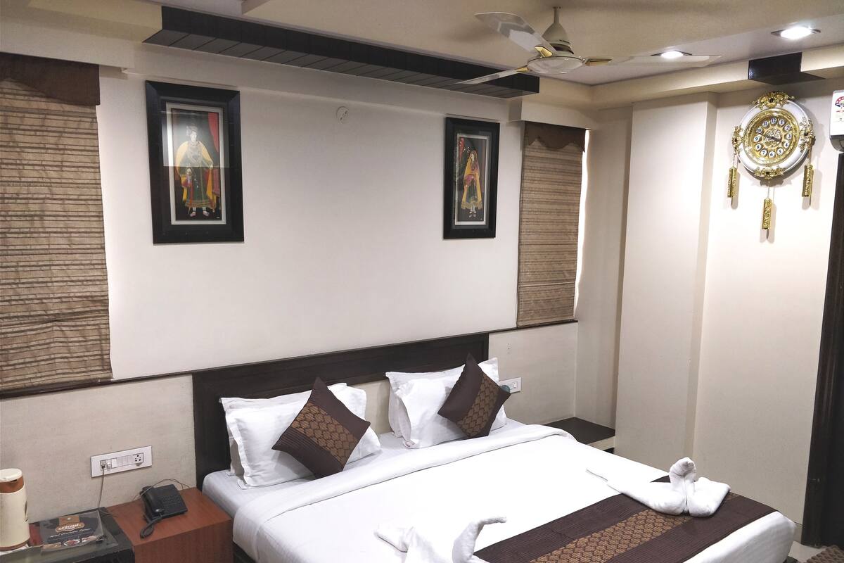 Quarto de hotel com cama bem arrumada, mesa de cabeceira com telefone e luminária, e relógio fixado na parede.
