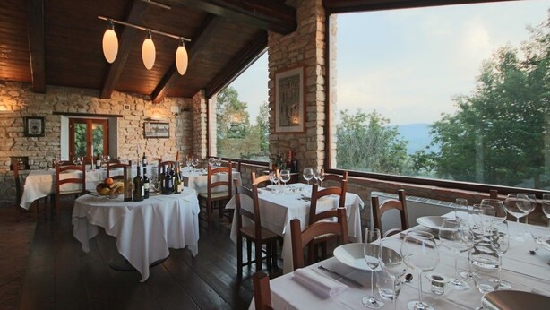 Un restaurante con paredes de piedra, vigas de madera y ventanas amplias que ofrecen vistas al exterior. Las mesas están preparadas con manteles blancos y copas de vino.