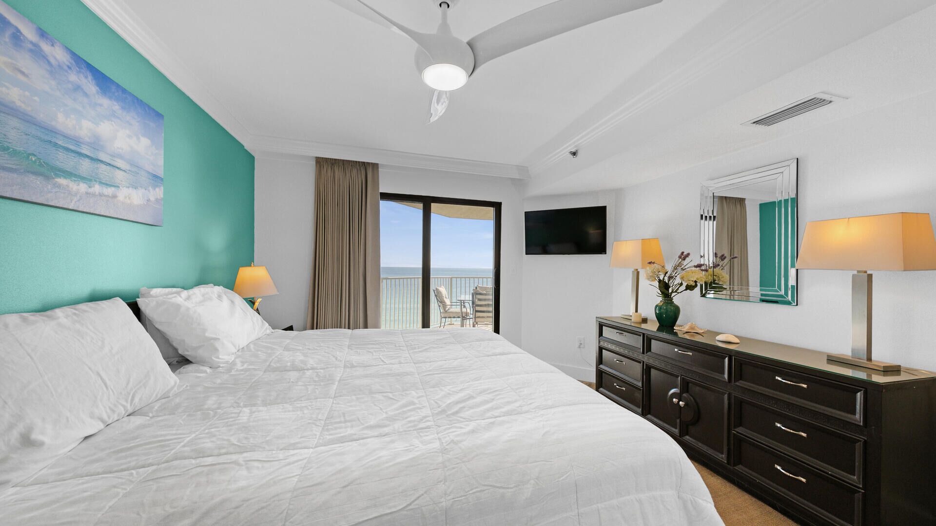 A hotel room with a large bed, a balcony with a view, a TV, and a dresser with lamps.