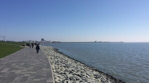 A paved walkway along a rocky shoreline with a clear view of the sea and distant buildings. - Pension Kruse (Büsum)