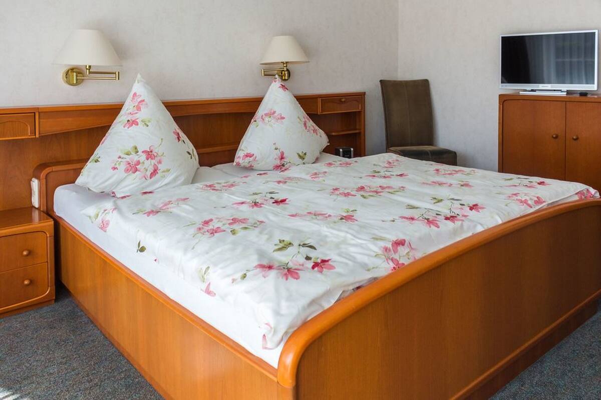A neatly made bed with floral bedding, a wooden headboard, bedside tables with lamps, a television on a wooden cabinet, and a chair.