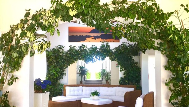 Un área de estar al aire libre acogedora, con un sofá curvo y sillones, rodeada de abundante vegetación y un mural con un atardecer.