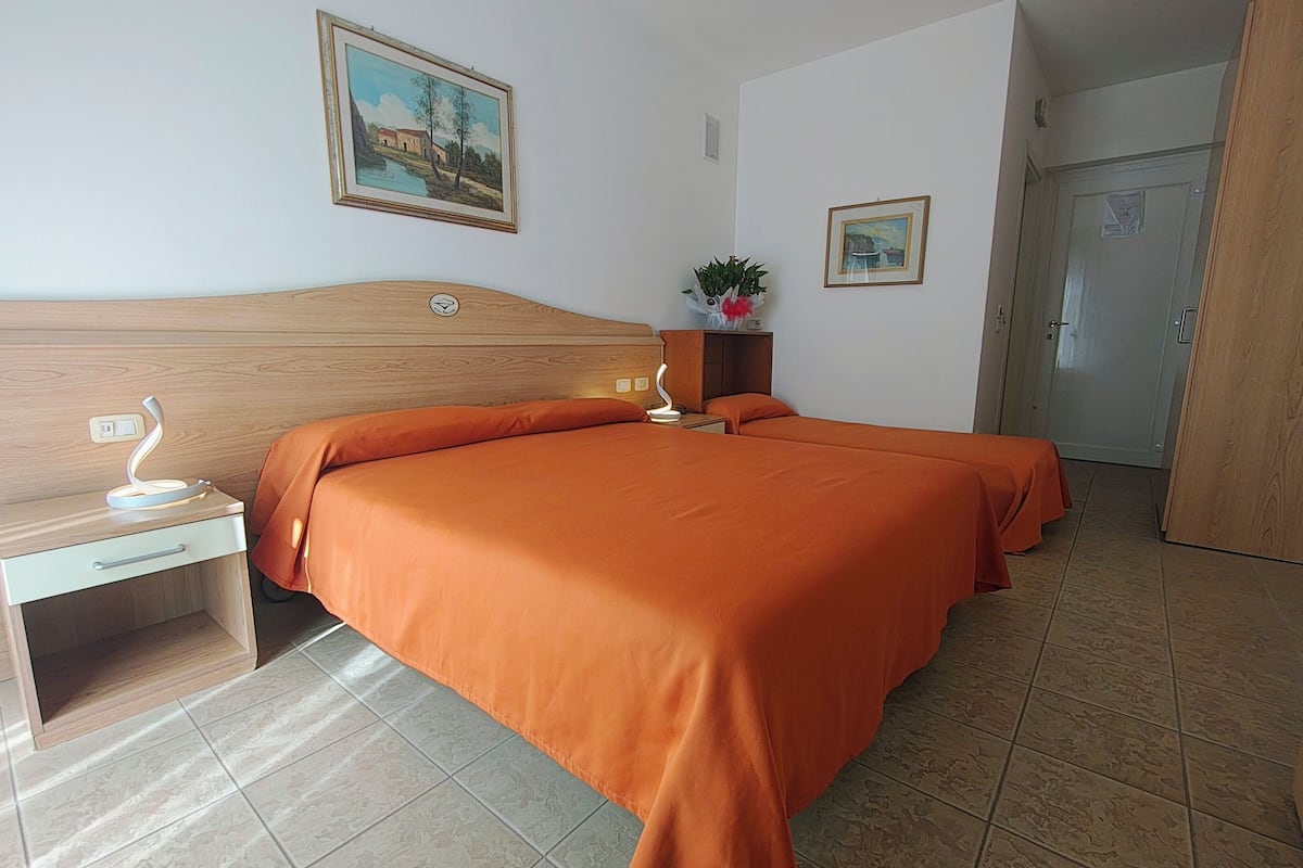 A hotel room with a wooden headboard, two beds with orange bedding, a nightstand with a lamp, a small table with a plant, and a door to the right.