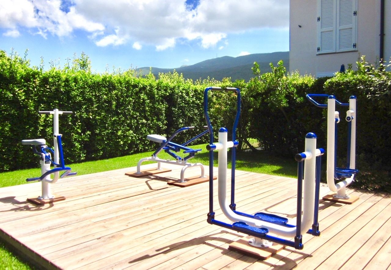 Buitenfitnessapparatuur op een houten terras, met uitzicht op een huis en bergen.