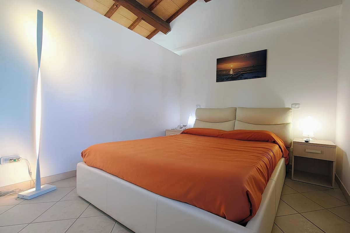 A bedroom with a bed, bedside tables, a nightstand, a painting, and a ceiling with wooden beams.