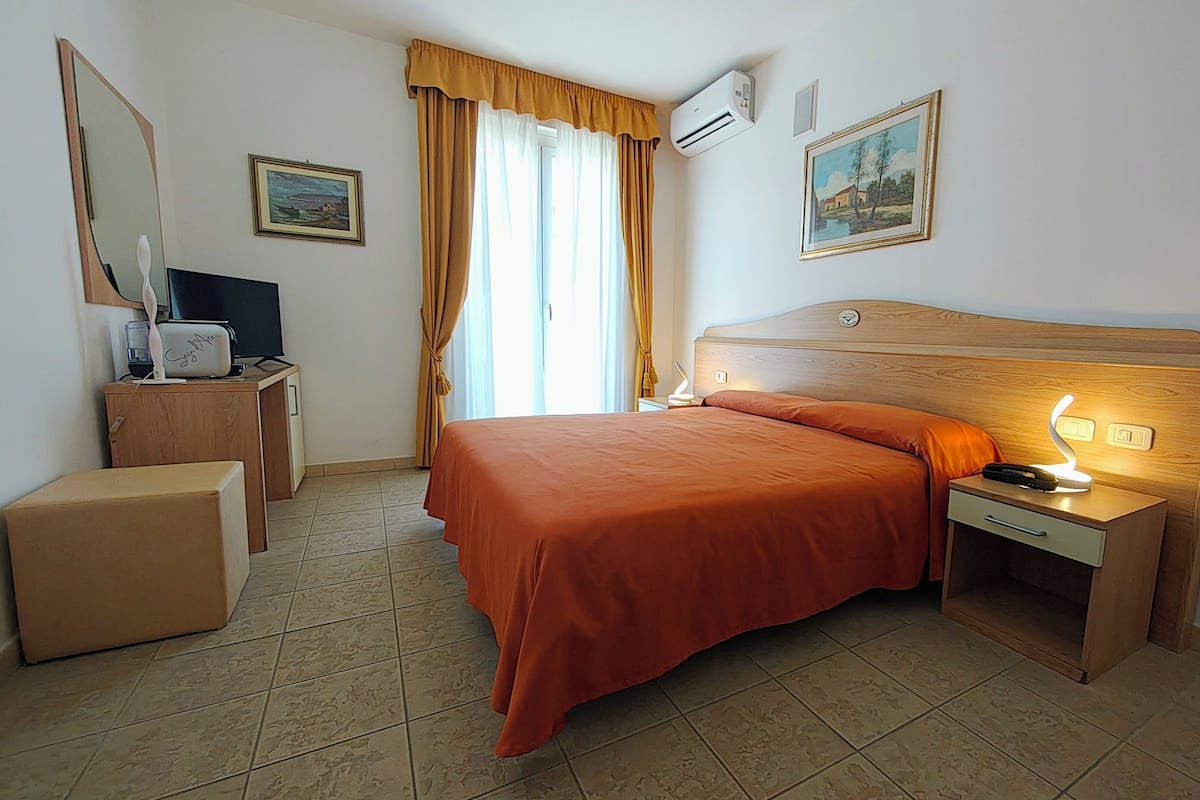 A hotel room with a bed, bedside table with a lamp, a dresser, a mirror, and two paintings on the wall.