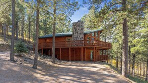 A log cabin with a stone chimney, wooden deck, and surrounded by tall pine trees. - The Reinerheim Cabin Centrally Located and Close To Lake Pactola (Rapid City)