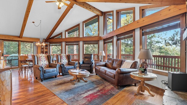 A spacious living room with wooden beams, large windows, leather sofas, and a stone fireplace. - The Reinerheim Cabin Centrally Located and Close To Lake Pactola (Rapid City)
