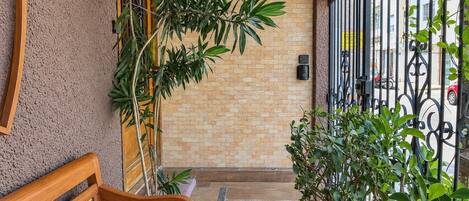 A wooden bench with a decorative backrest, a potted plant, and a metal gate in a courtyard with a brick wall and a tiled floor.