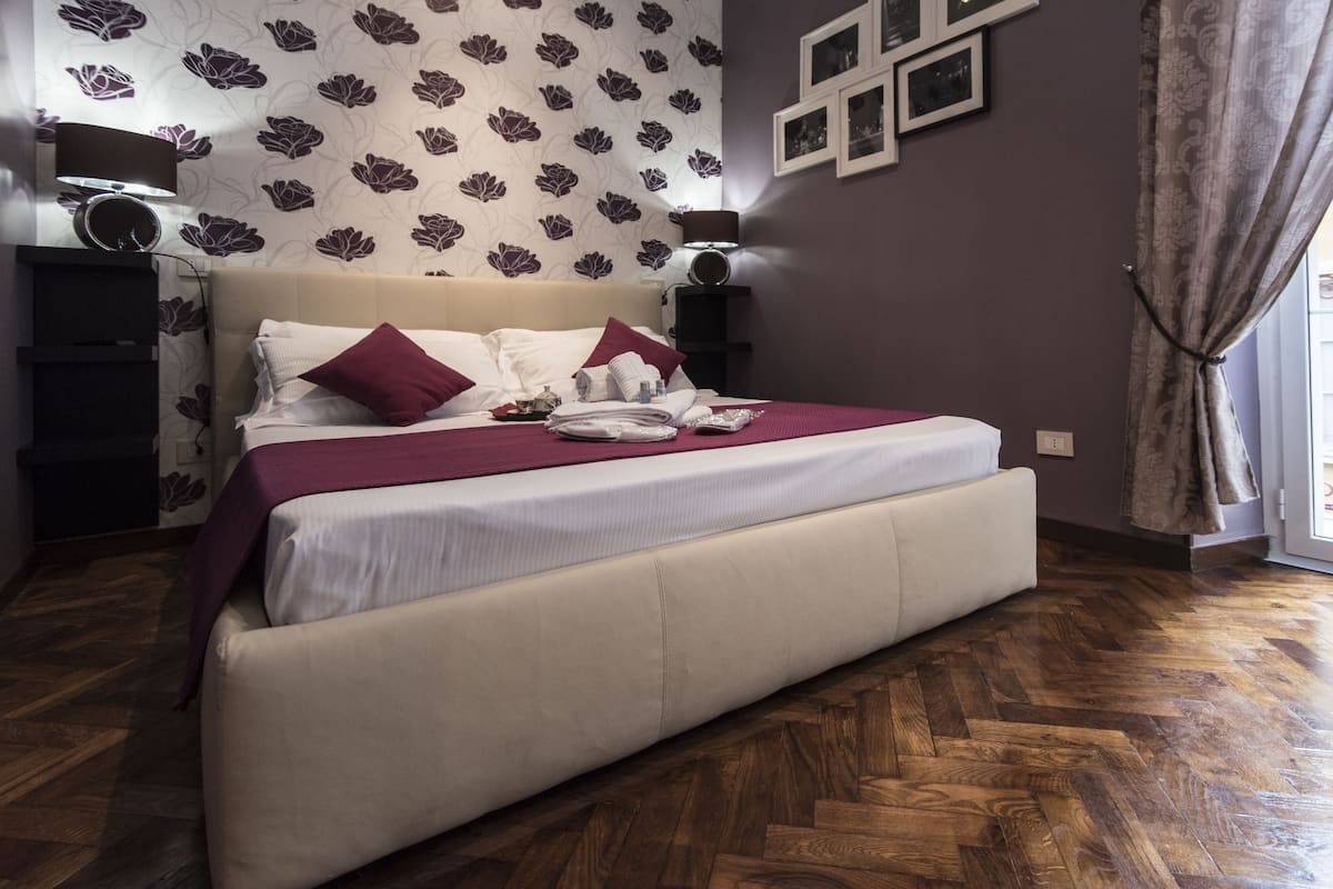 A large bed with a headboard, a bedside table, a lamp, and a wall with framed pictures.