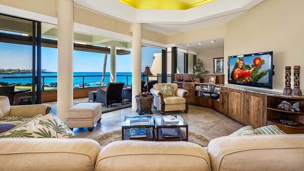 A spacious living room with a large TV, comfortable seating, a view of the ocean, and a ceiling fan. - 2BR ocean and sunset views in Mauna Lani point (Kamuela)