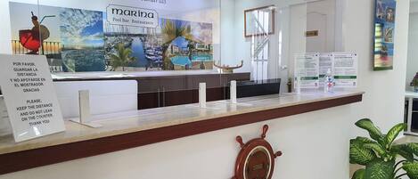 A reception desk at Marina Apartments with a view of the marina, a ship's wheel decoration, and informational posters.