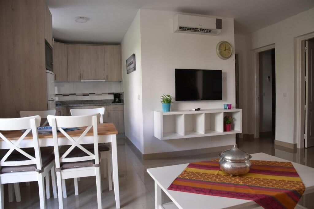 A modern living room with a flat-screen TV, a dining table with chairs, and a kitchen area in the background.