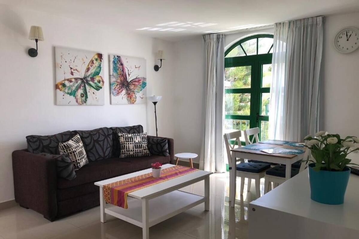 A living room with a sofa, coffee table, and a dining area with a table and chairs. There are butterfly paintings on the wall, a clock, and a window with curtains.