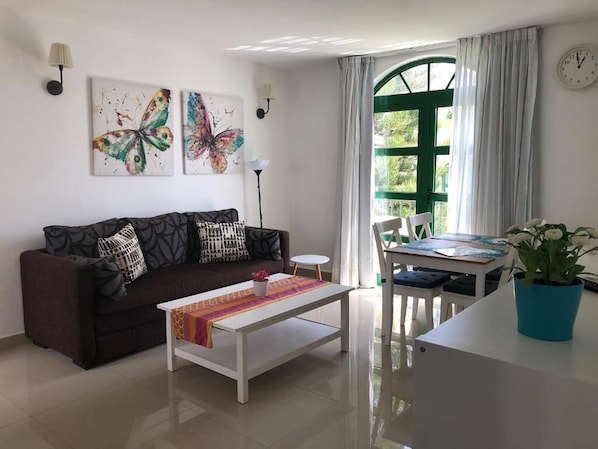 A living room with a sofa, coffee table, and a dining area with a table and chairs. There is a window with curtains, a wall clock, and artwork on the wall. - Billy's Puerto de Mogán Apartments by LIVVO (Mogan)