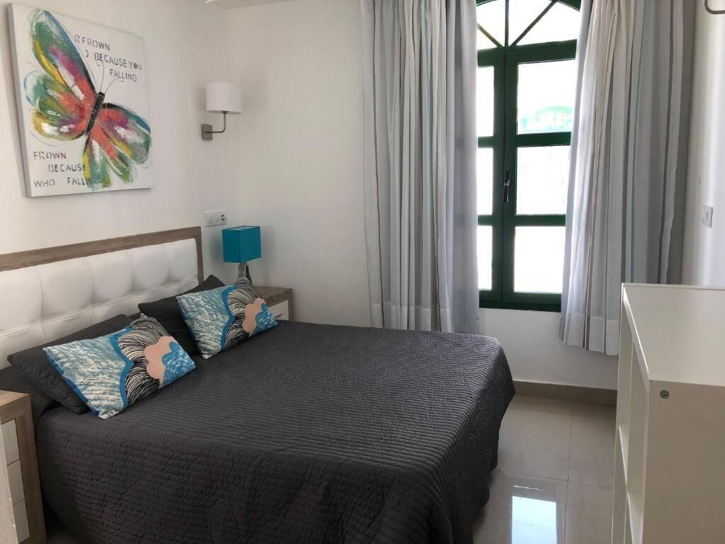 Un dormitorio con cama, una lámpara de pared, una ventana con cortinas y un cuadro en la pared.