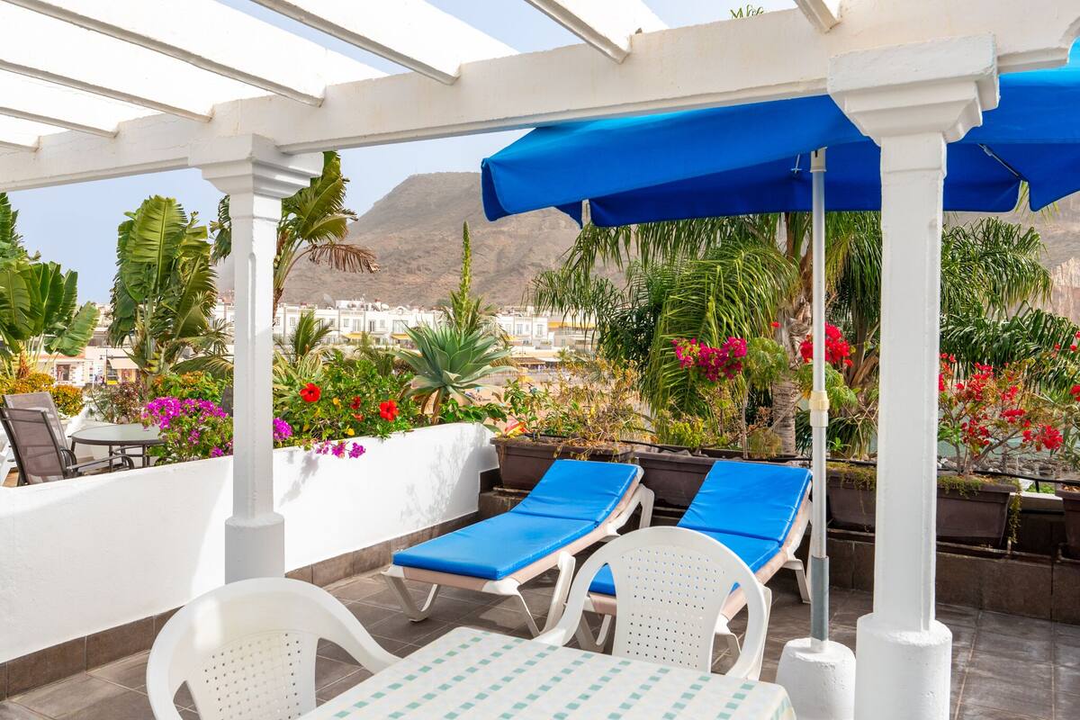 A covered outdoor seating area with lounge chairs and a table, surrounded by potted plants and flowers, overlooking a coastal town.