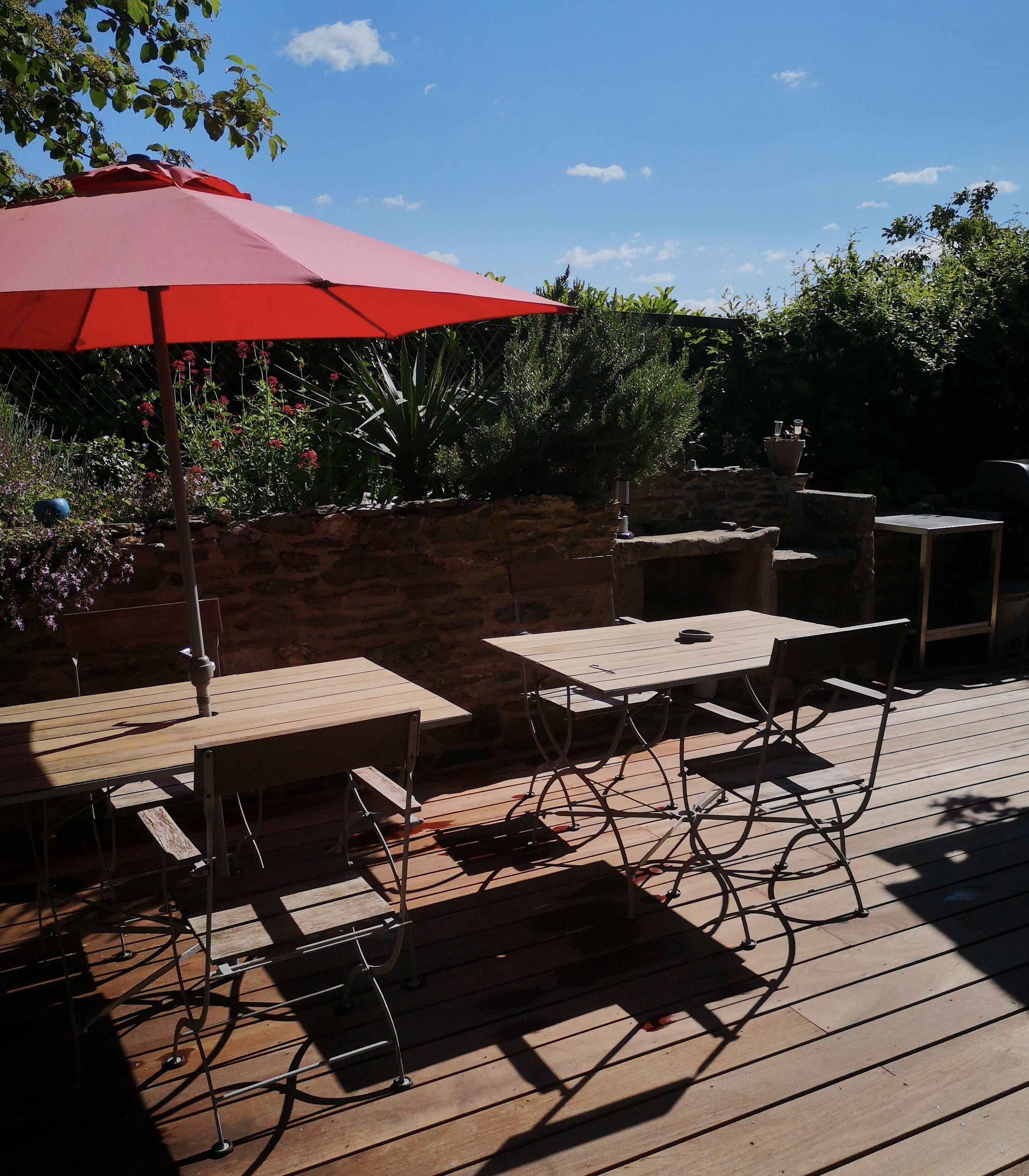 Un patio al aire libre con mesas y sillas de madera, un paraguas rojo y plantas en macetas.