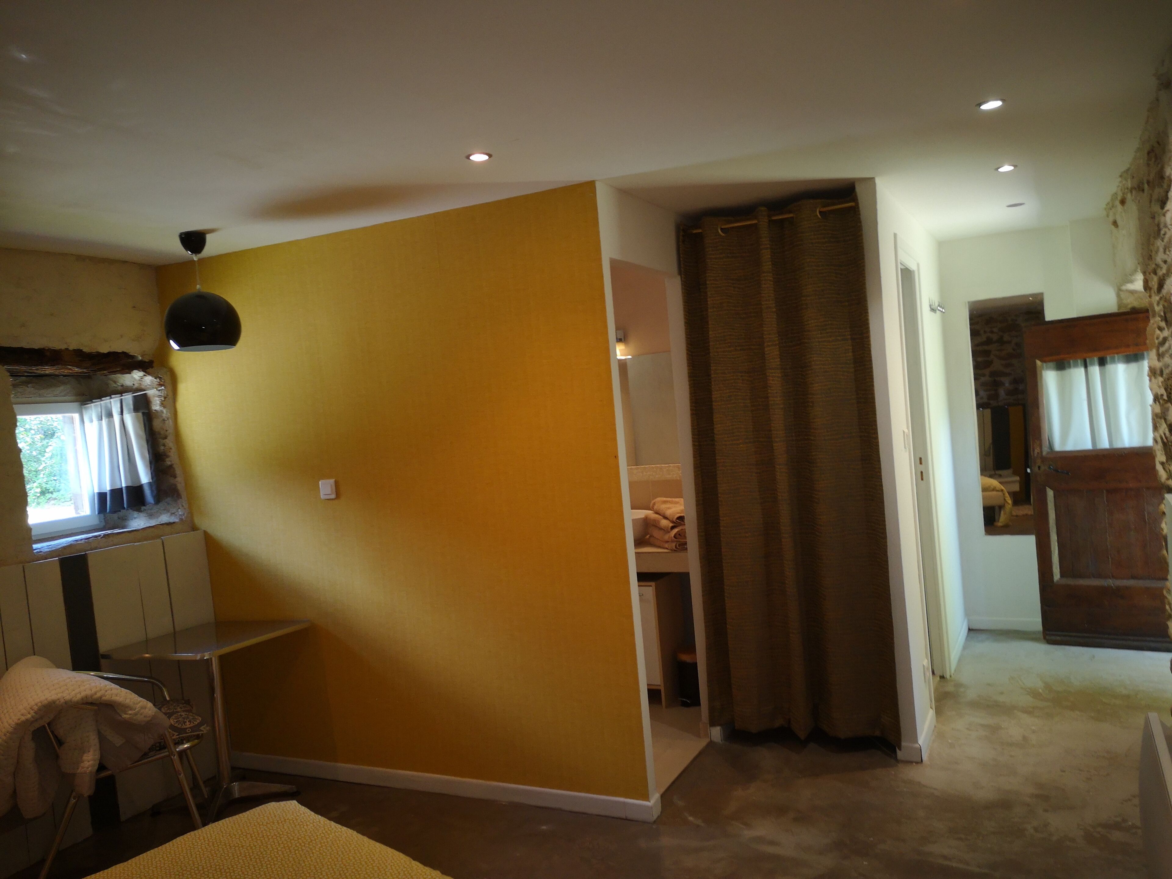 A room with a yellow wall, a window with curtains, a bed, and a bathroom visible through an open door.