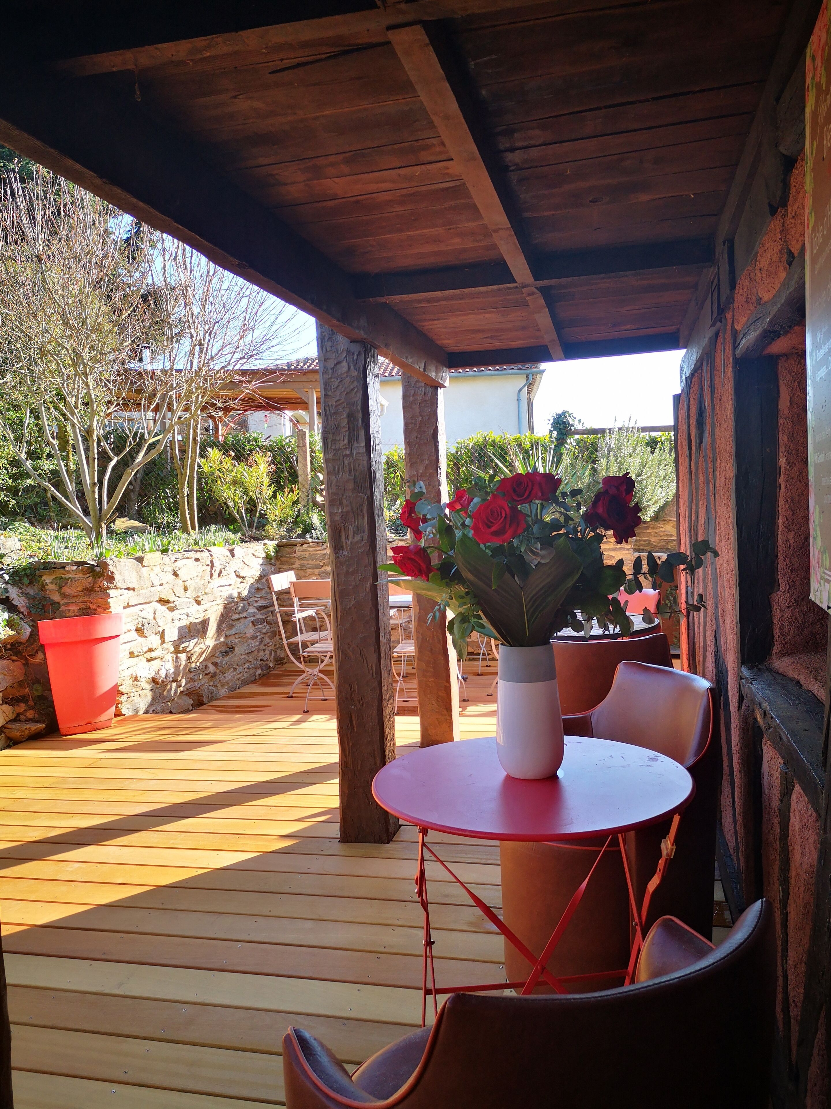 Un patio al aire libre con piso de madera, una mesa redonda y sillas rojas. Hay plantas en macetas y un muro de piedra al fondo.