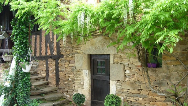 A stone building with a wooden door and steps leading up to it. There are potted plants and hanging baskets around the entrance.
