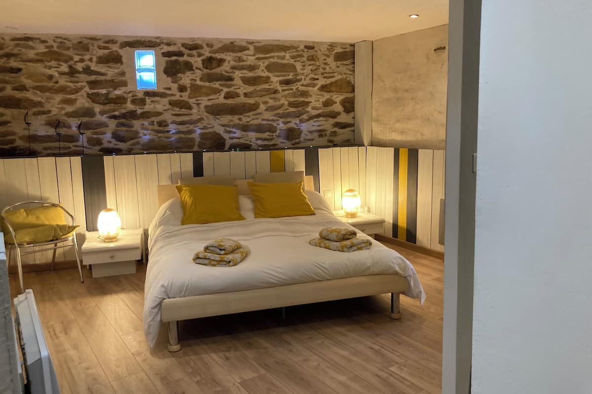Une chambre avec un mur en pierre, un lit avec une literie blanche et des coussins jaunes, deux lampes de chevet et un fauteuil.
