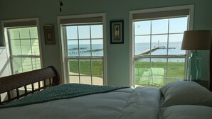 A bedroom with a large bed, two windows with a view of the sea, a bench, and a lamp.
