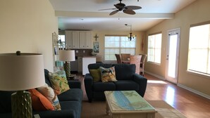 A spacious living room with a dark blue sofa, a coffee table, and a lamp. There is a kitchen area in the background with white cabinets and a dining table with chairs.
