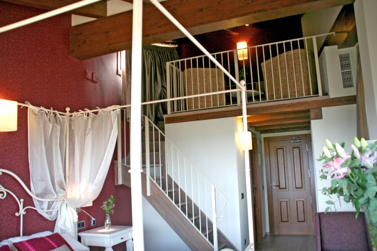 Un dormitorio con cama, mesita de noche, ventana con cortinas, una escalera y una puerta.
