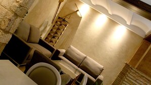 A cozy, well-lit lounge area with a sofa, chairs, and a small table, featuring a stone wall and a wooden ceiling.