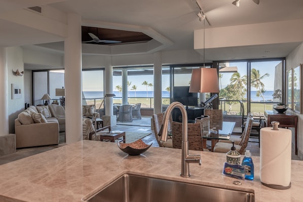 A modern kitchen with a large island, a dining area with wicker furniture, and a view of the ocean. - 2BR Mauna Lani Point with stunning Ocean Views! (Kamuela)