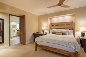 A bedroom with a wooden bed, two bedside tables with lamps, a ceiling fan, and a glass door leading to a bathroom.