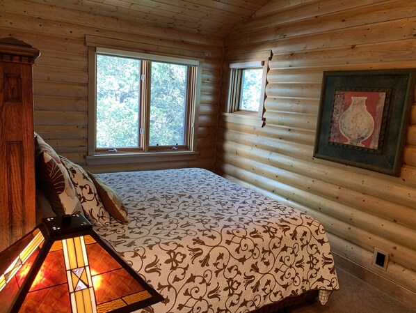 A log cabin bedroom with a bed, a nightstand, a lamp, and a window with a view of trees. - Luxury Mountain Home. Hot Tub, 6 Bedrooms, 5.5 Bath, Theater, Steam Rm, Sauna (Sundance)
