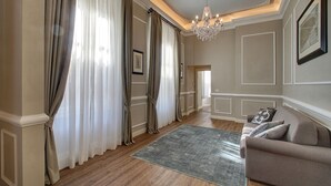 Ein Zimmer mit Holzboden, beige Wänden, einem beigen Sofa, einer Kristallleuchtre und einem großen Fenster mit Vorhängen.