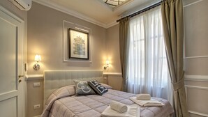 Ein ordentlich gemachtes Bett mit einer gesteppten Decke, zwei Kissen und einem gefalteten Handtuch. Darüber befinden sich zwei Wandleuchten und ein gerahmtes Bild.