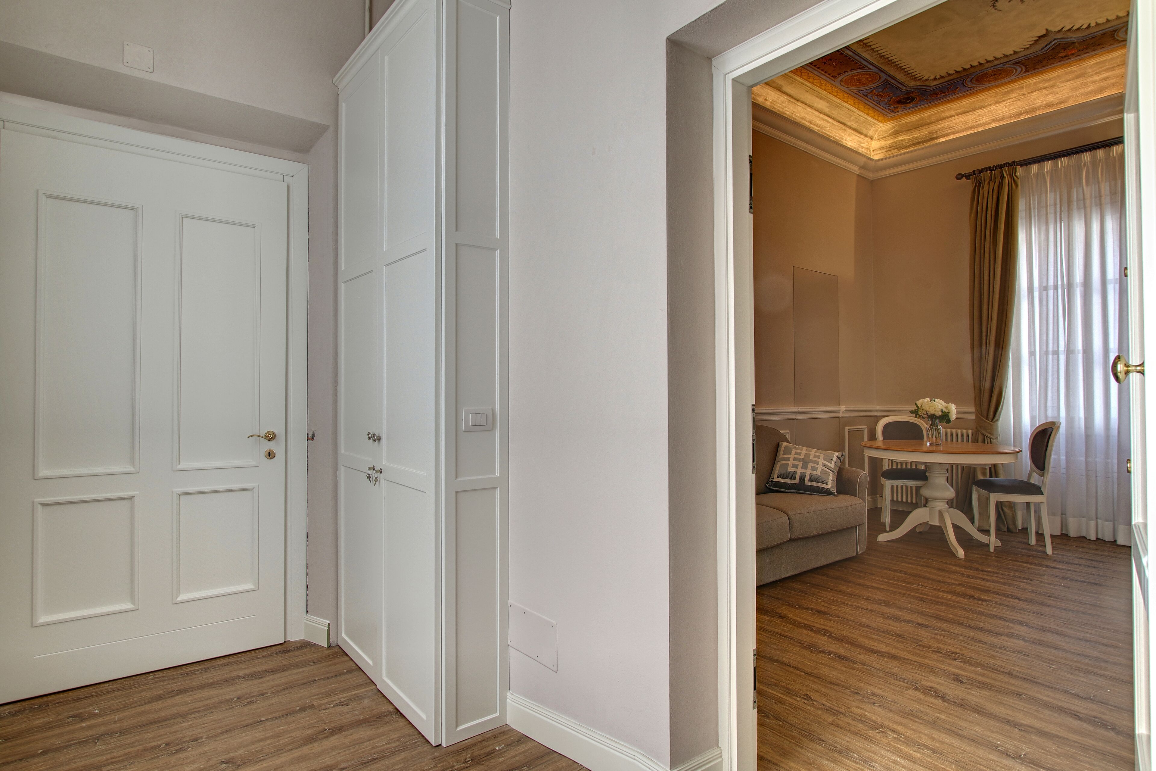 Een kamer met een houten vloer, witte muren en een witte deur. Er is een zithoek met een tafel, stoelen en een bank.