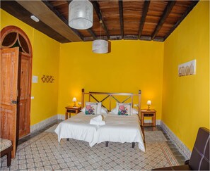 A room with a bed, two bedside tables, a wooden wardrobe, a patterned floor, and a yellow wall.