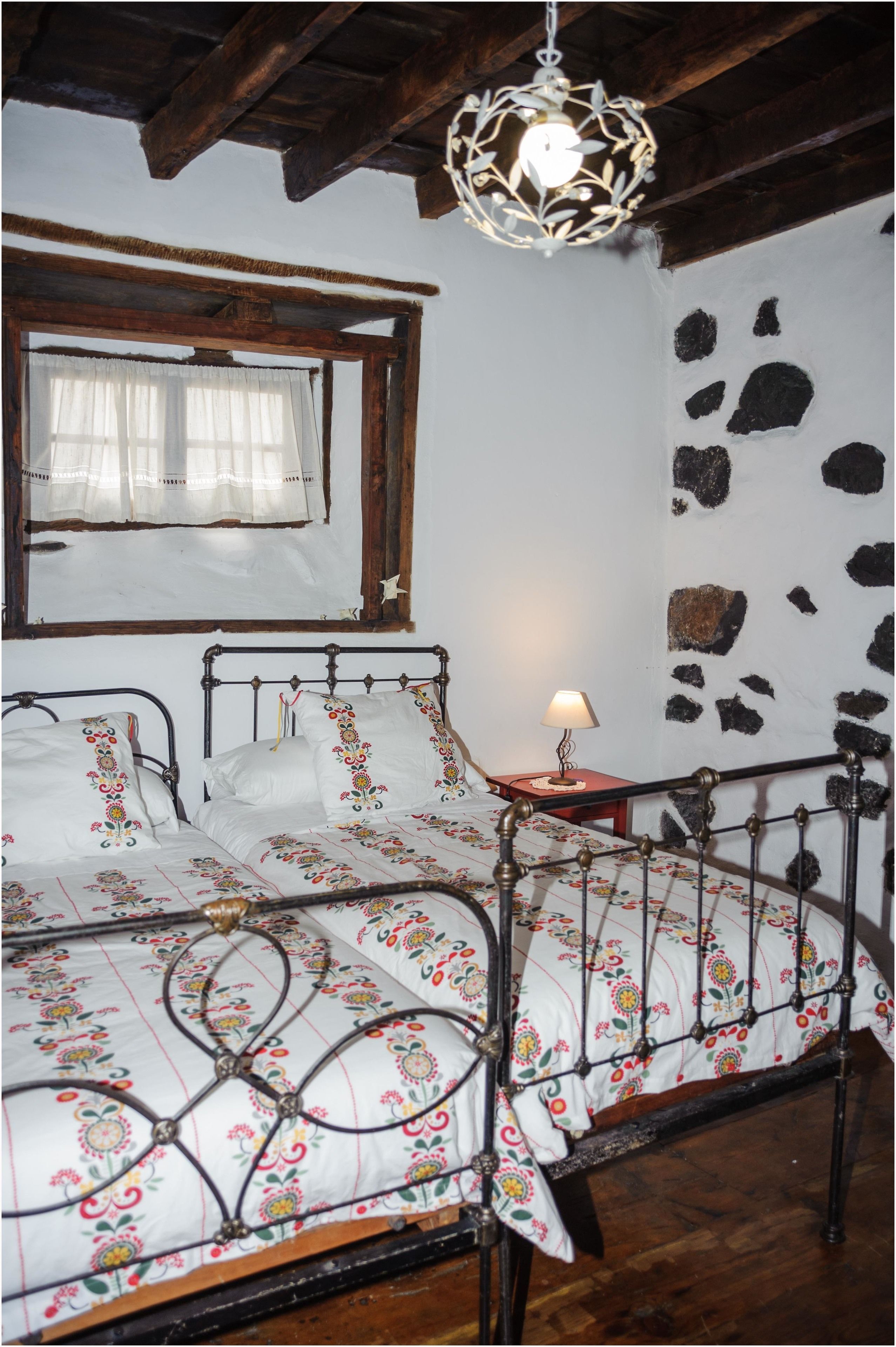 A rustic bedroom with a metal bed frame, floral bedding, a wooden ceiling, a stone wall, and a hanging light fixture.