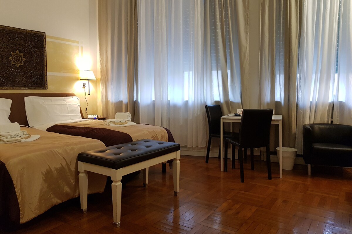 A hotel room with two beds, a dining table, chairs, a bench, and a painting on the wall.