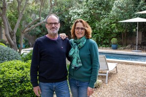 Un hombre y una mujer están de pie en un jardín con una piscina y un banco al fondo.