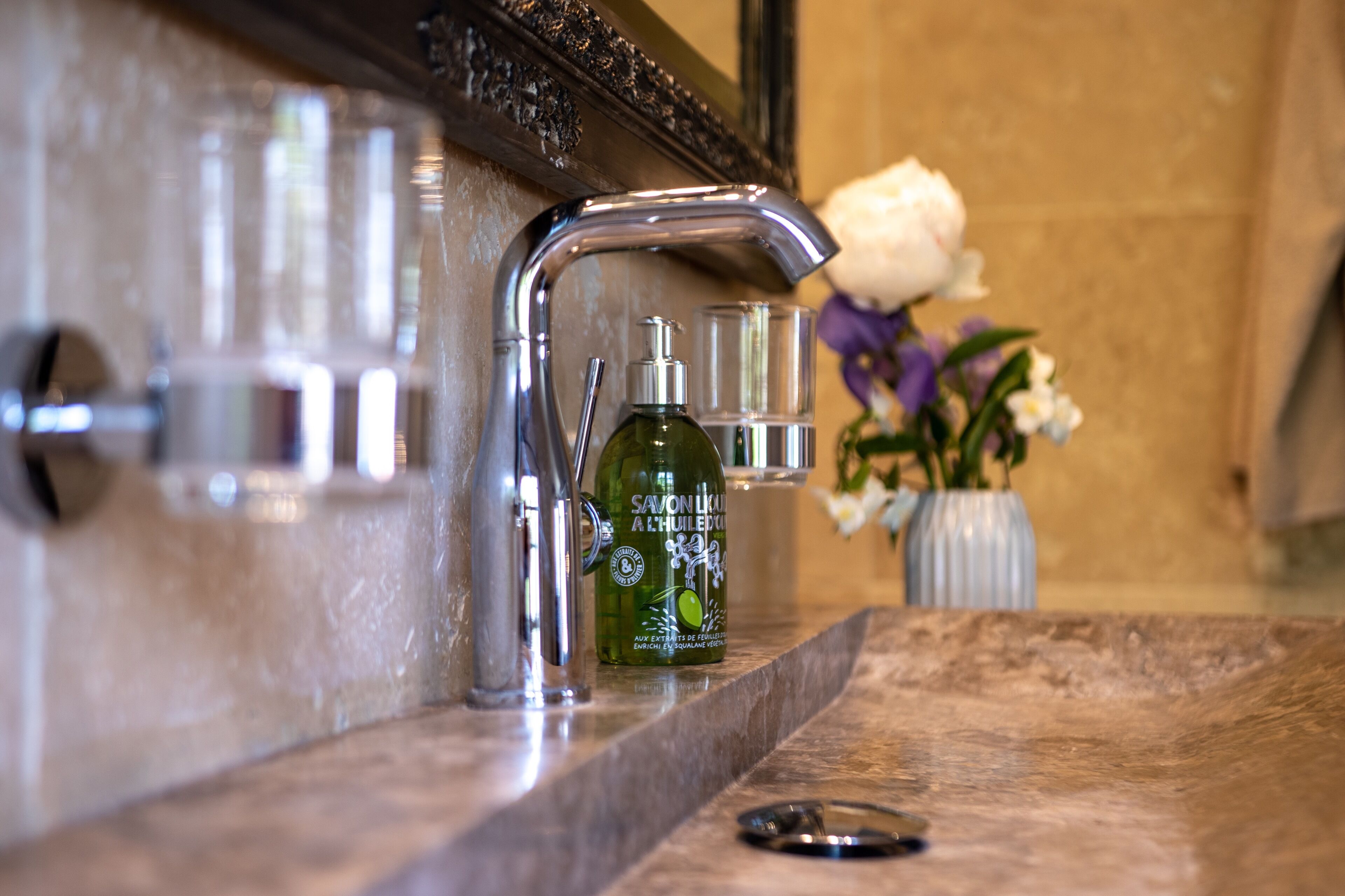 A bathroom vanity with a mirror, a faucet, a soap dispenser, and a vase with flowers.